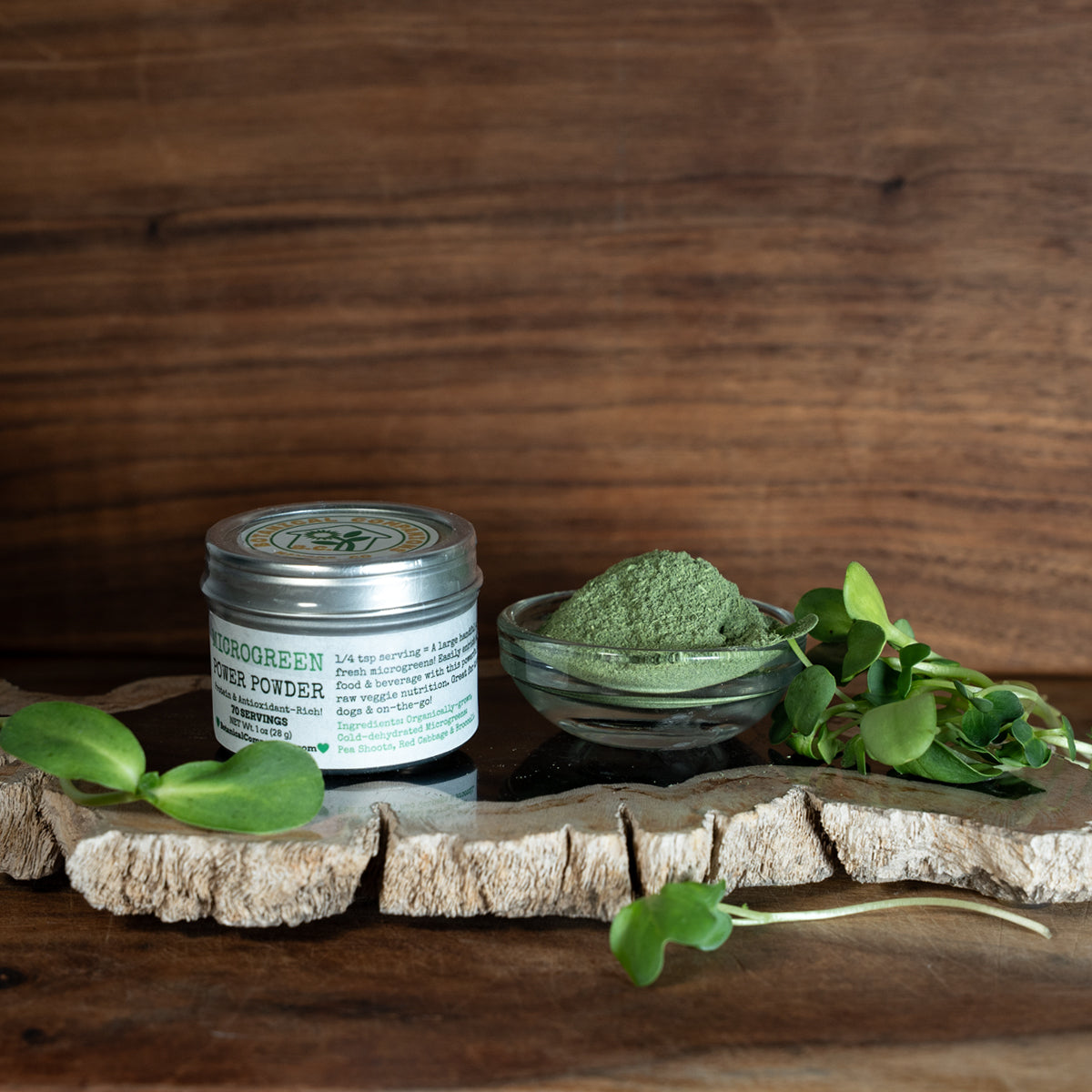 A tin labeled 'Botanical Companions’ and ‘Microgreen Power Powder' with product info including 1 ounce, 70 servings, and a 1/4 teaspoon serving size being equivalent to a handful of fresh microgreens with an ingredient list of organically grown, cold-dehydrated microgreens: Pea Shoots, Red Cabbage, Brocolli, and is placed next to a small glass bowl filled with pure green microgreen powder, placed on a petrified wood surface with fresh microgreen sprouts scattered around.